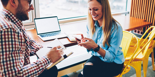 a man and a woman are sitting at a desk facing each other. On the desk is an open laptop